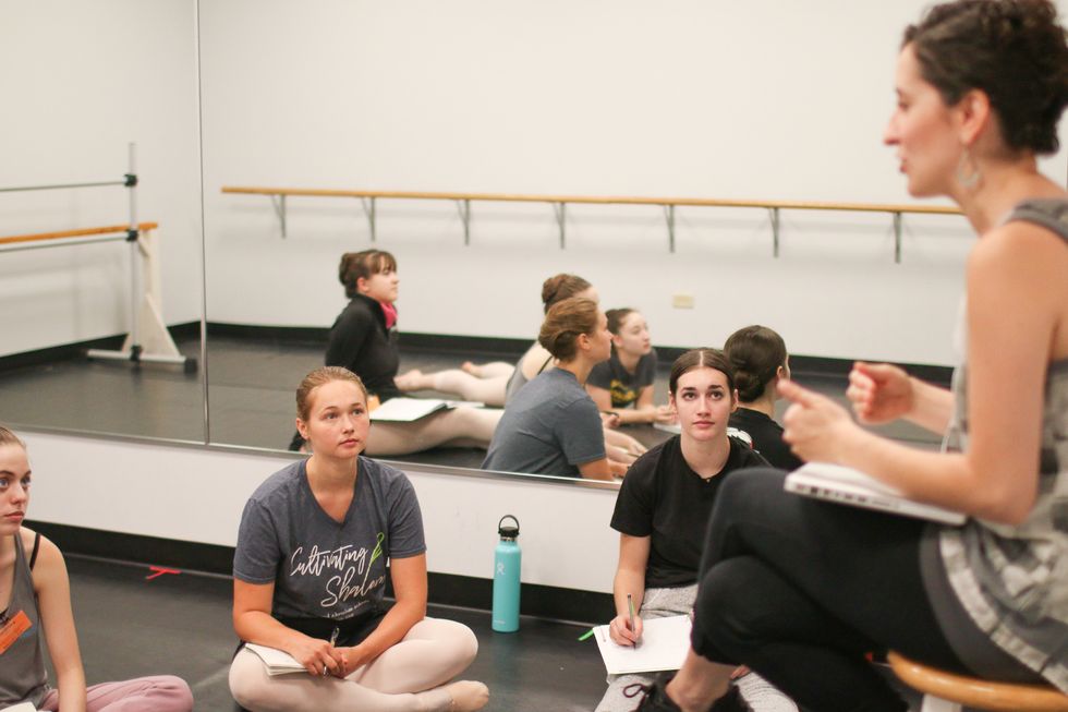 Three young dancers sit crosslegged on a dance studio floor, wearing casual T-shirts and pink tights and shoes. A faculty member sits on a chair in front of them leading a discussion with a laptop on her lap; she wears dark leggings and a gray, loose-fitting tank top.