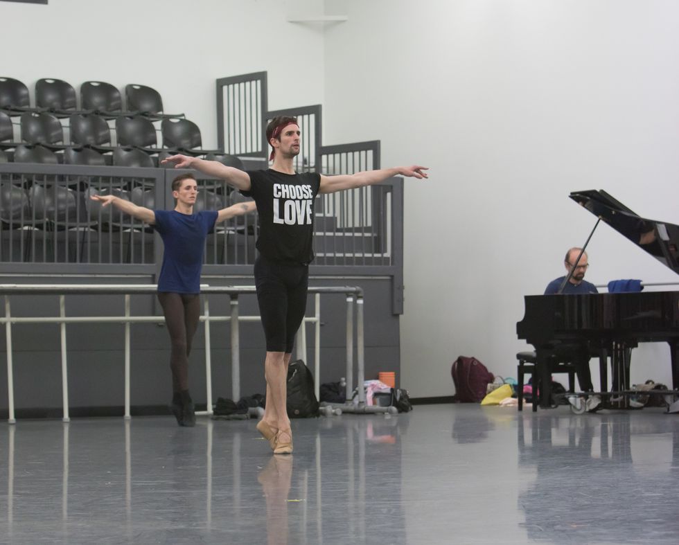Two male dancers in practice clothes stand in sous-sus with their arms out in second position