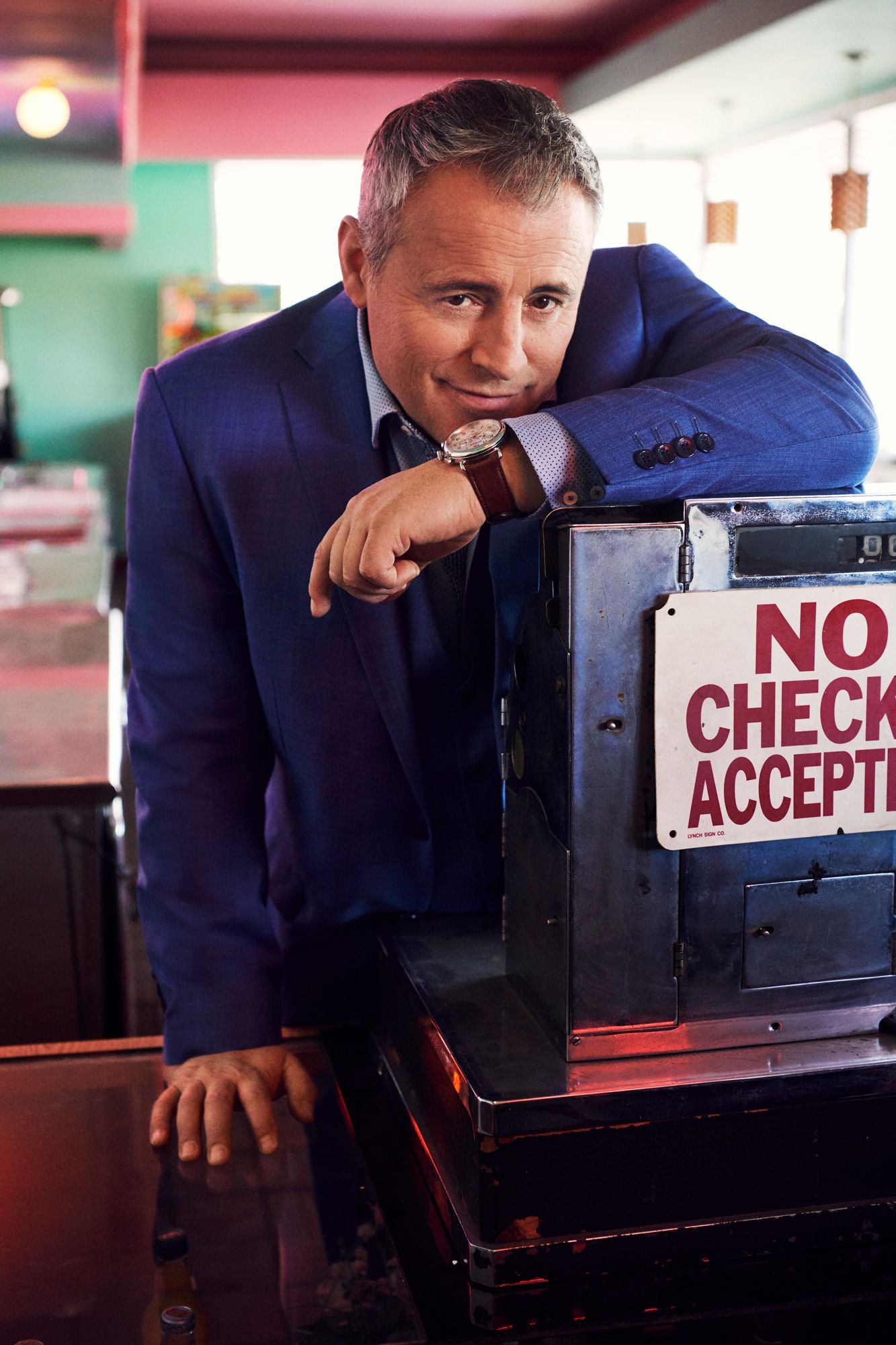 Matt LeBlanc at a vintage cash register looking into the camera wearing a blue suit.