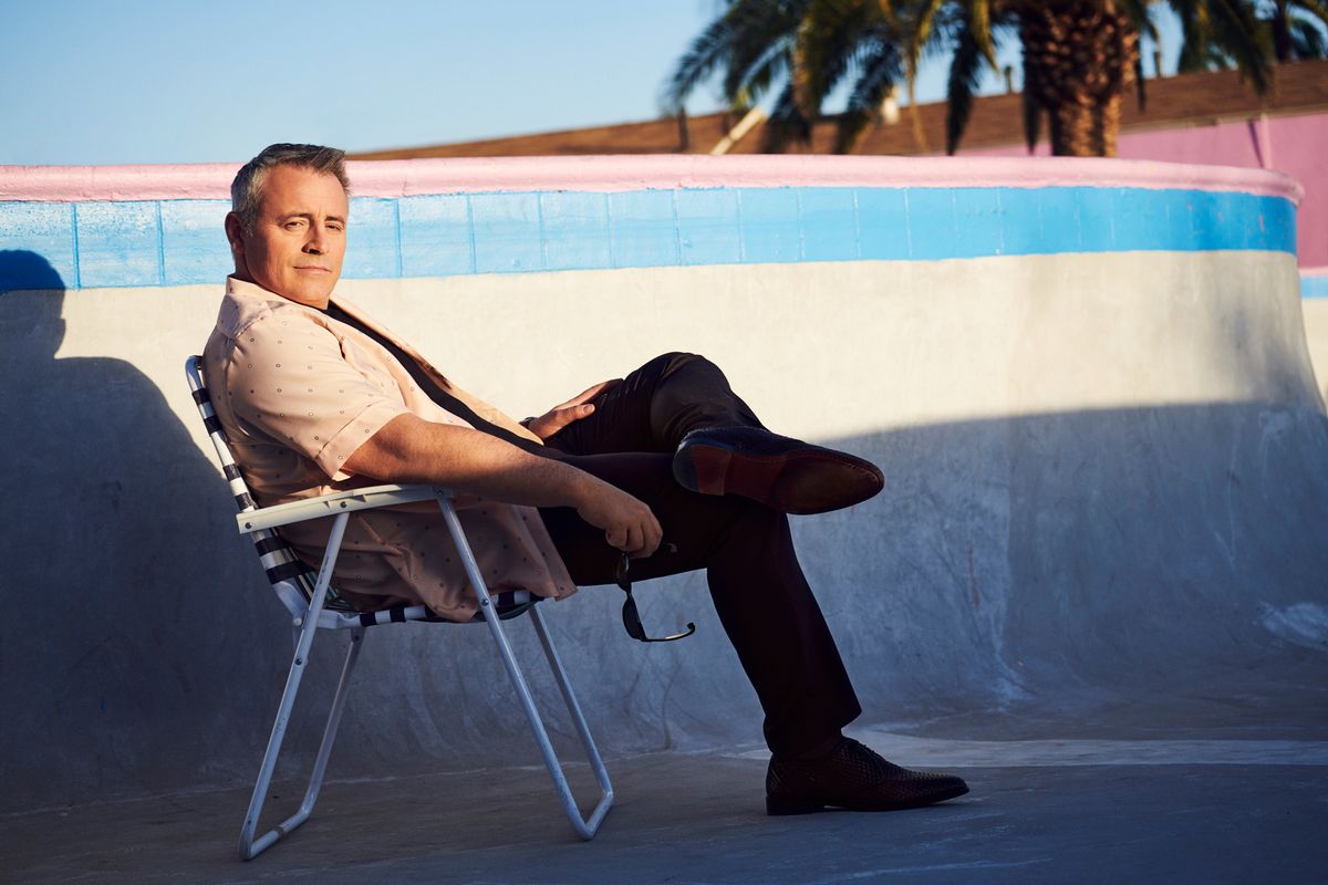 Matt LeBlanc sitting in an empty swimming pool wearing a vintage short-sleeve collar shirt.