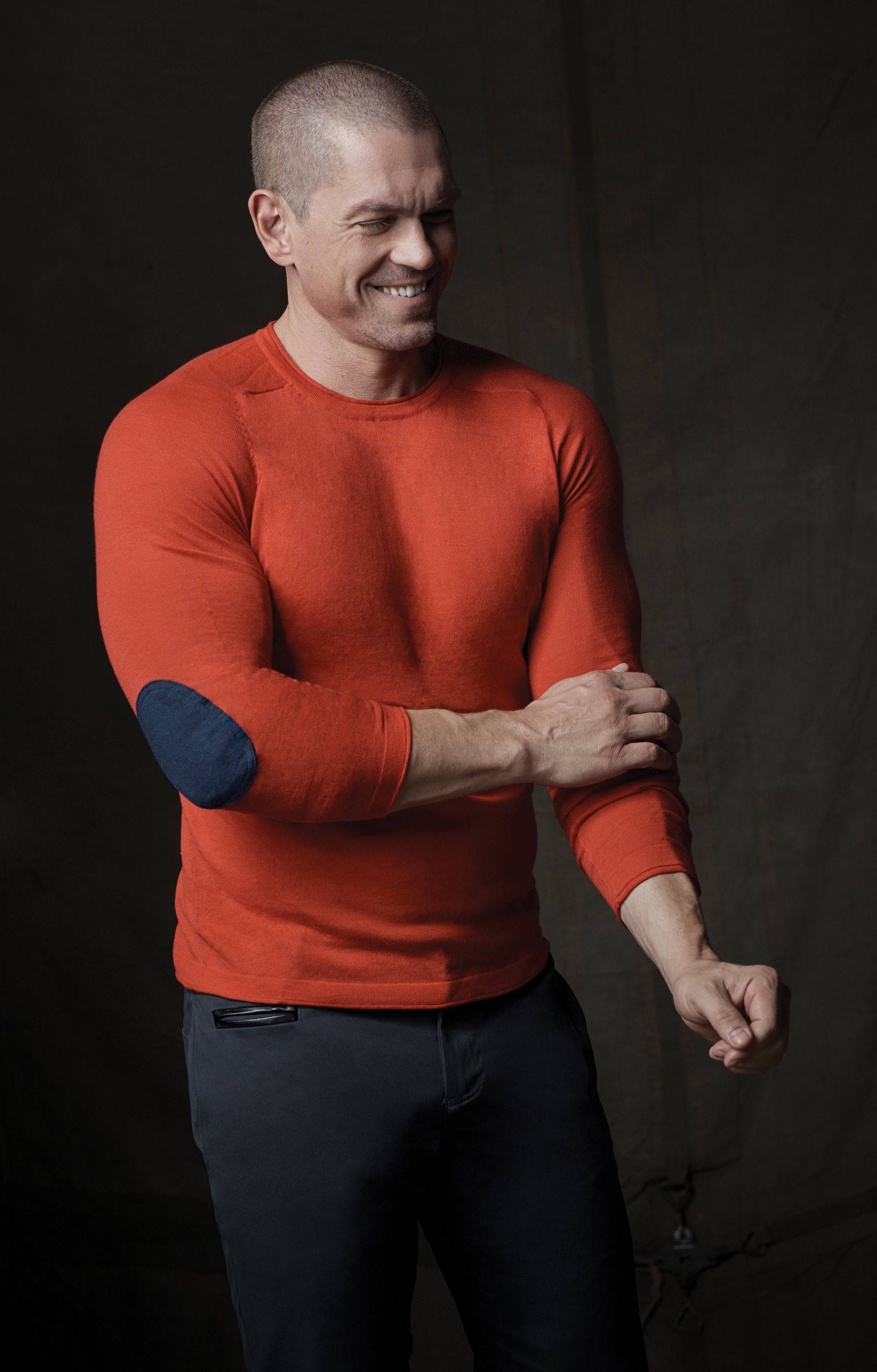 Actor Steve Howey laughing and wearing an orange sweater.
