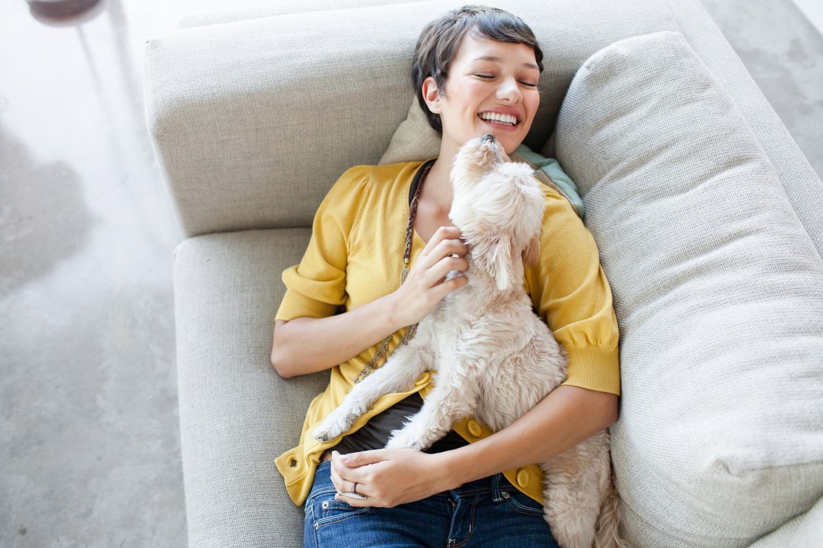 A woman in yellow with a pixie cut cuddles a small white dog on a white couch