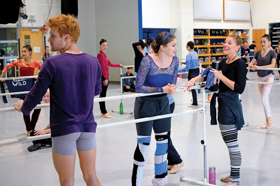 Morgan stands with a group of dancers at the barre before class. She smiles and leans on the barre as another dancer is talking.