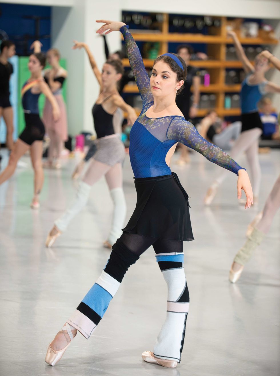 Morgan, in a blue leotard, black tights and skirt and color blocked legwarmers stands in a tendu front in class, with a serious expression on her face.