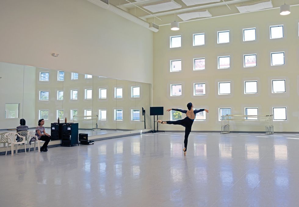 Morgan is seen from behind in an arabesque on pointe. Lopez sits in a white plastic chair in front of the mirror, watching her.