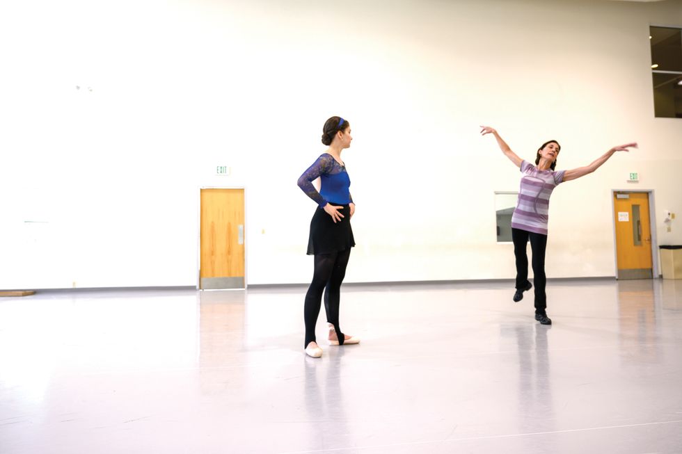 Morgan stands with her hands on her hips while Lopez, dressed in black exercise pants and a purple striped athletic t-shirt, demonstrates a sequence from Firebird.