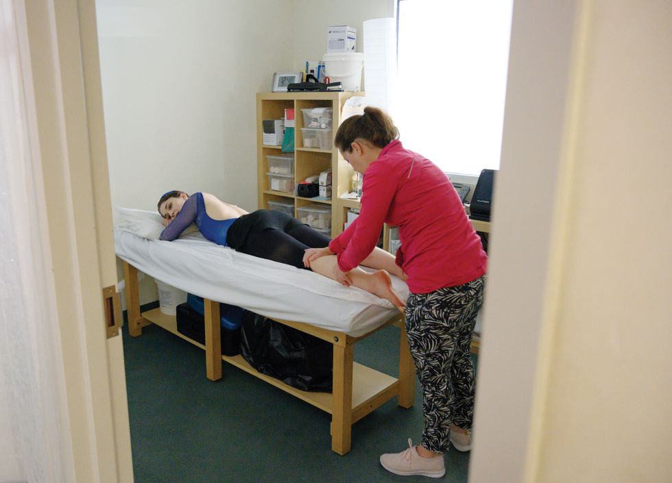 Morgan lies face down on a massage table. A body worker in a red top and patterned pants stands behind her, working on her calf.