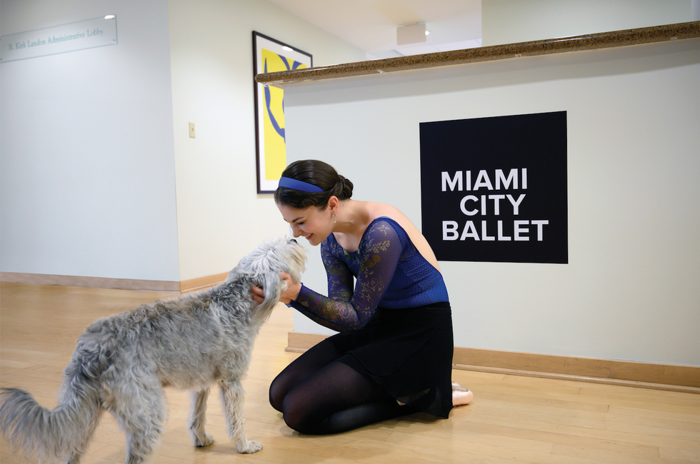 Morgan kneels on the floor with her hands on a scruffy gray dog. Behind her is a counter with the words Miami City Ballet printed.