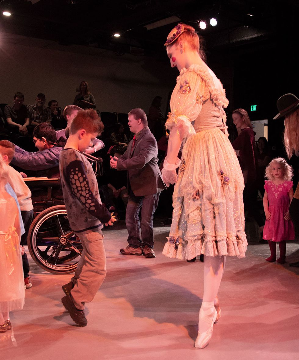 A ballerina demonstrates standing on relev\u00e9 to a young boy. They're surrounding by other children and adults practicing dance moves.