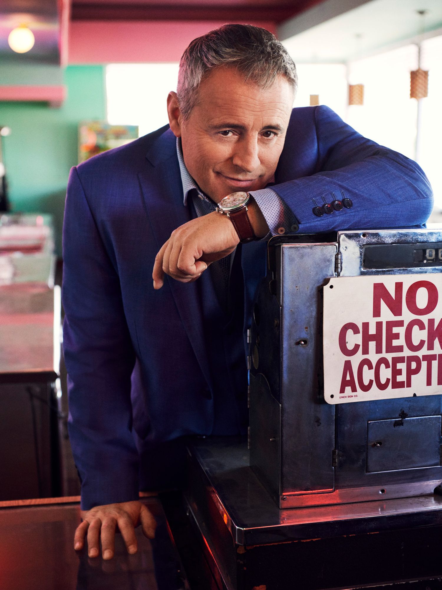 Matt LeBlanc at a vintage cash register looking into the camera wearing a blue suit.