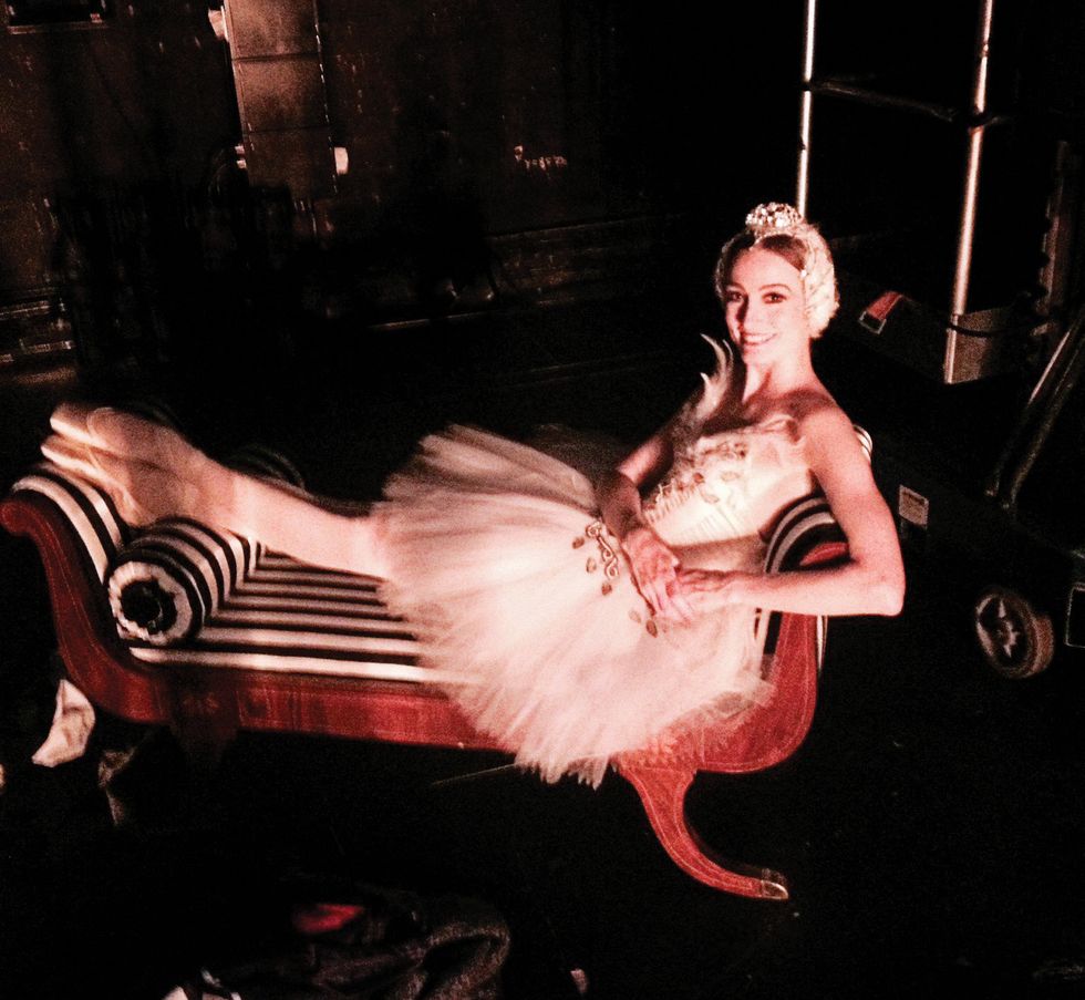 A grainy photograph of Julie Kent in a white swan tutu and headpiece, lounging on a striped chaise lounge with her hands on one hip and her feet crossed at the ankle.