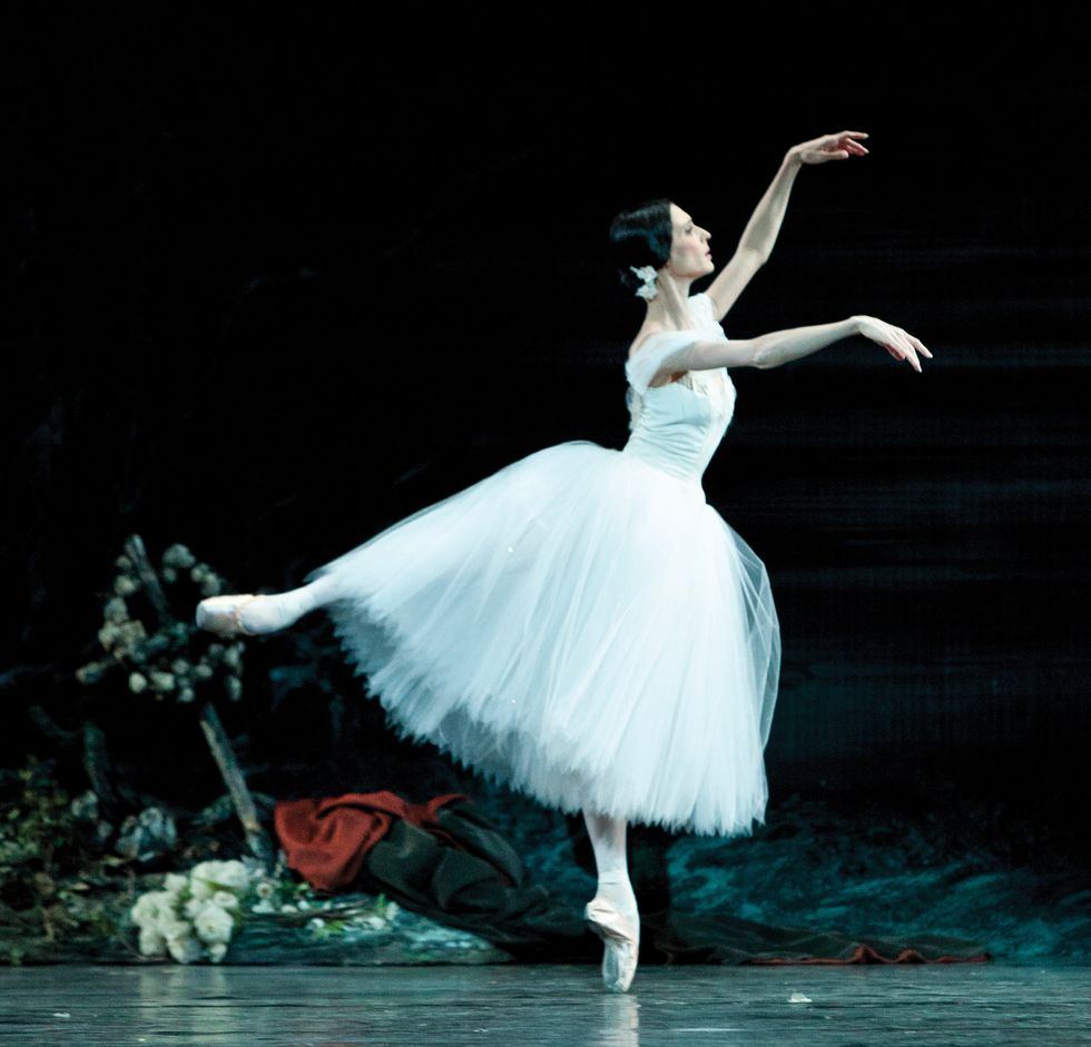 Hodgkinson, in a white tutu, in an arabesque on pointe with her arms in fourth position.