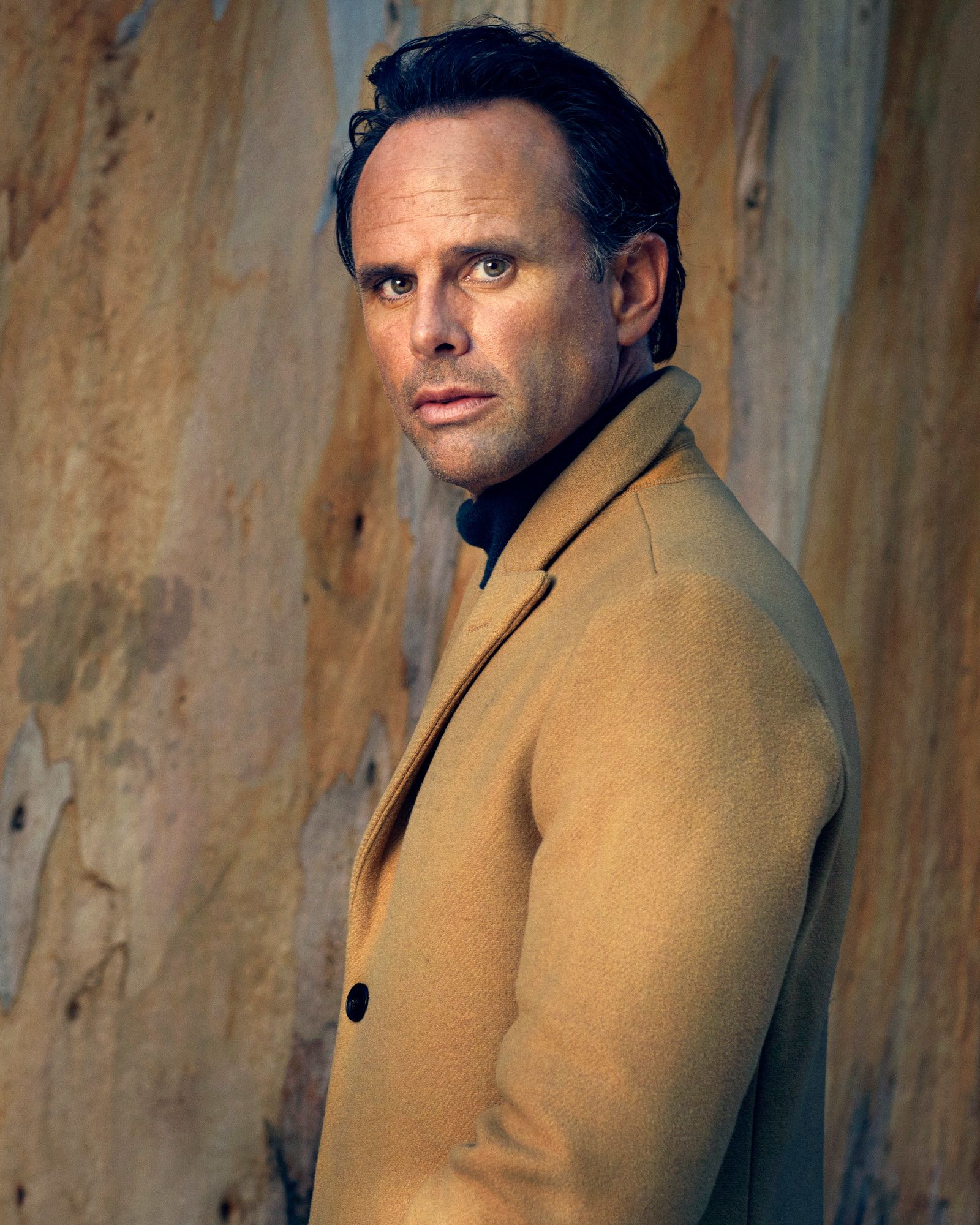 Walton Goggins looking sideways into the camera wearing a camel coat.