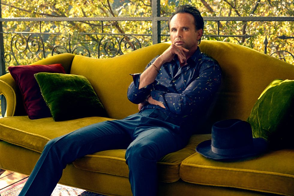 Walton Goggins sitting on a mustard color velvet sofa staring into the camera.