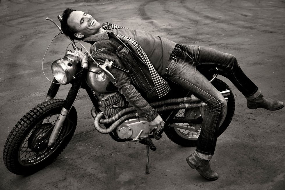Black and white image of Walton Goggins reclining on a vintage motor cycle.