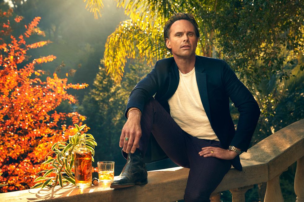 Walton Goggins sitting on a stone balustrade, sun backlit with fall colors, a glass of whiskey beside him.