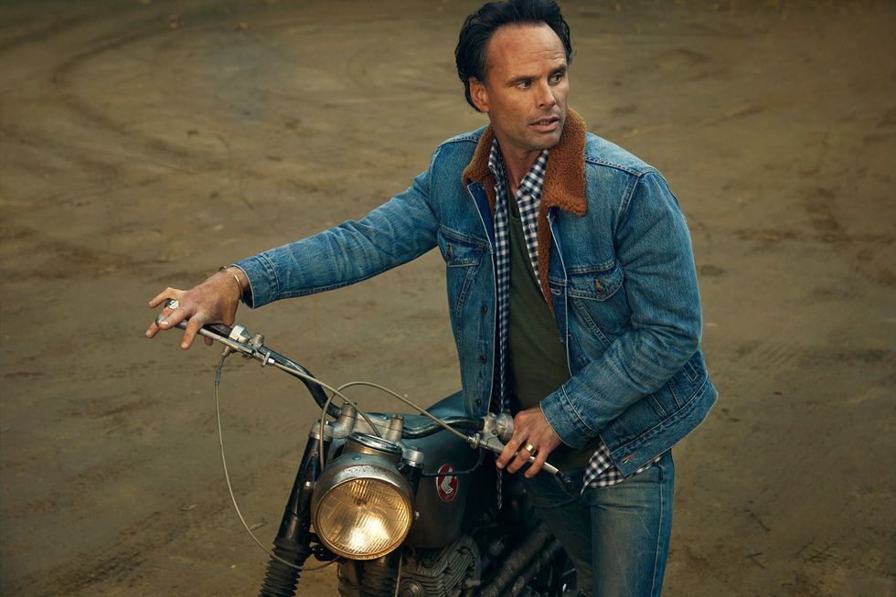 Walton Goggins in a seated pose on a vintage motor cycle, looking sideways.
