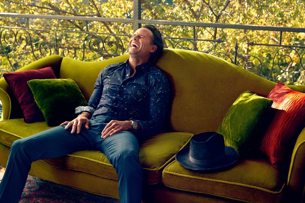 Walton Goggins lounging and laughing on a mustard color velvet sofa.
