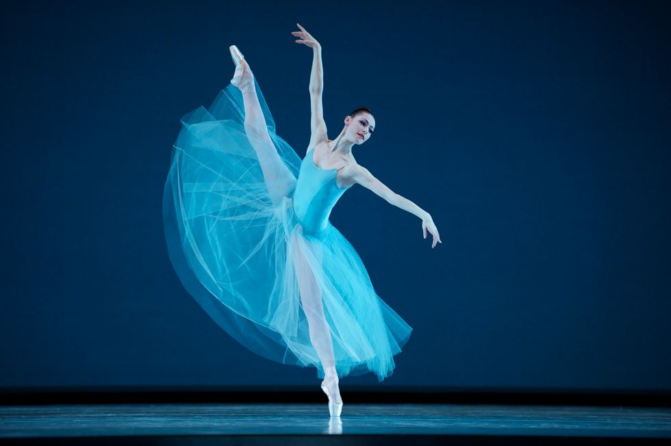 Sylvie, in a long light blue tutu and leotard, stands off-center on pointe with one leg extended high in the air. She looks over her left arm.