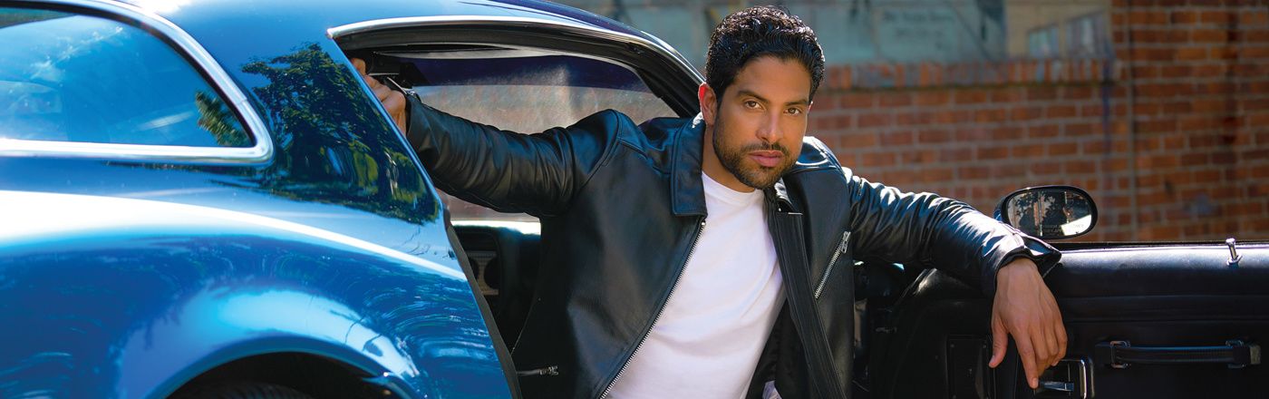 Adam Rodriquez in an opened leather jacket and white t-shirt in the passenger seat of a car with the door open