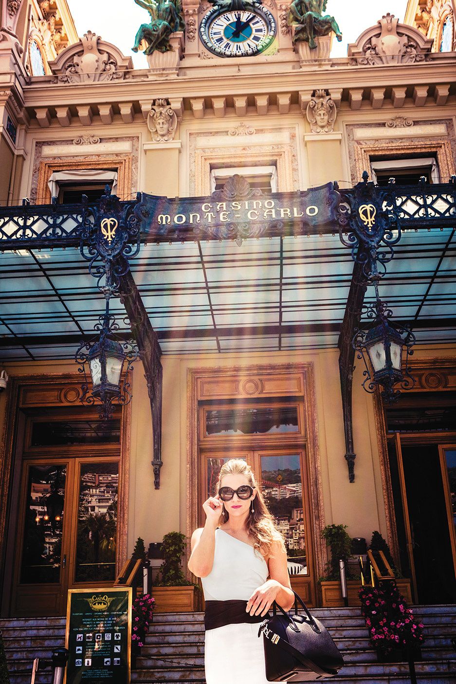 AJ Cook in large sunglasses and a white dress in front of Casino Monte Carlo