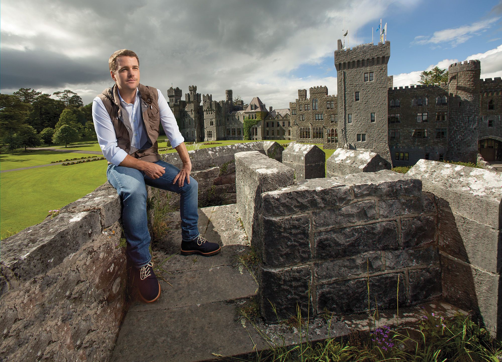 Chris O'Donnell sitting on stone wall with Ashford Castle behind him.