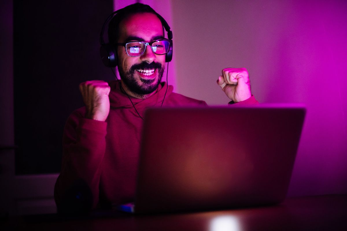 A man in glasses with headphones cheers at his laptop