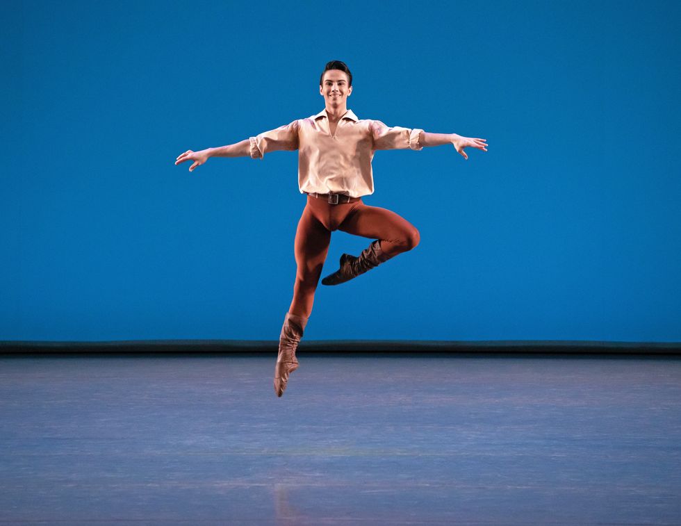 Mejia onstage in a light brown blouse and brown leggings and boots jumping in the air against a blue background. He's smiling at the audience.