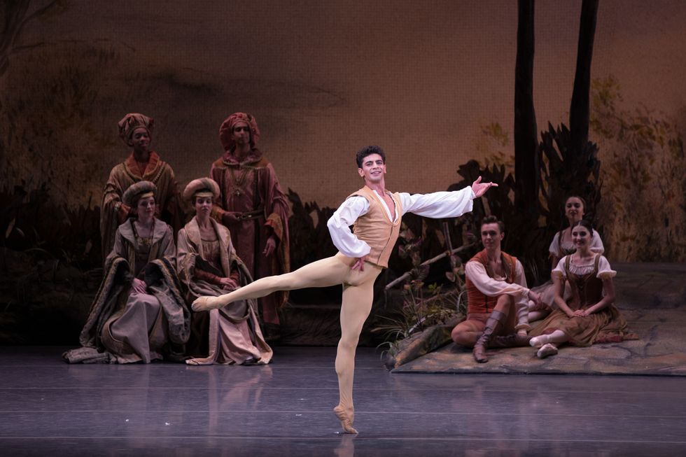 Mkrtchyan in brown tights and vest and a white blouse standing onstage in an arabesque with his hand on his hip. A group of dancers dressed as villagers and royalty sit behind him.