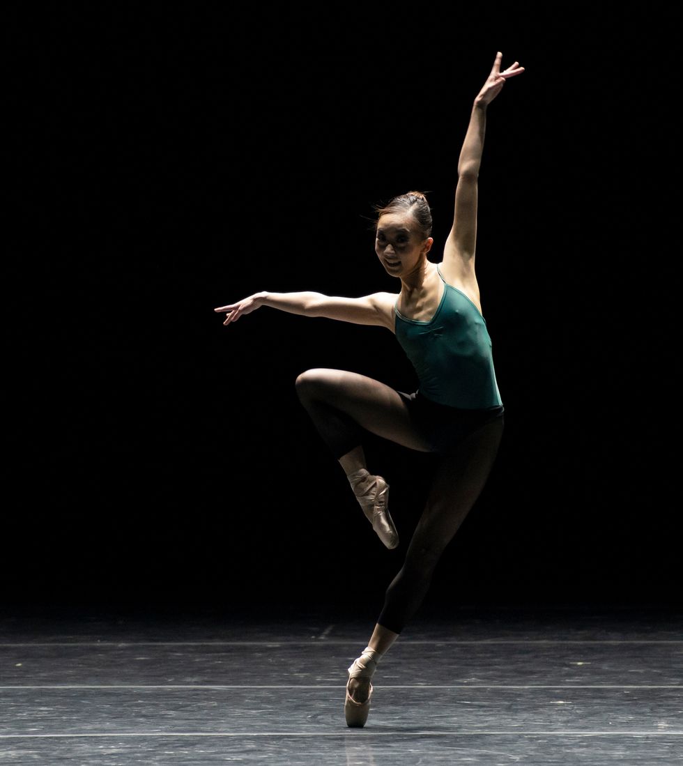 Oga, in a green leotard and black tights and pointe shoes, is onstage in an off-kilter pass\u00e9 with her arms in a jazzy extension.