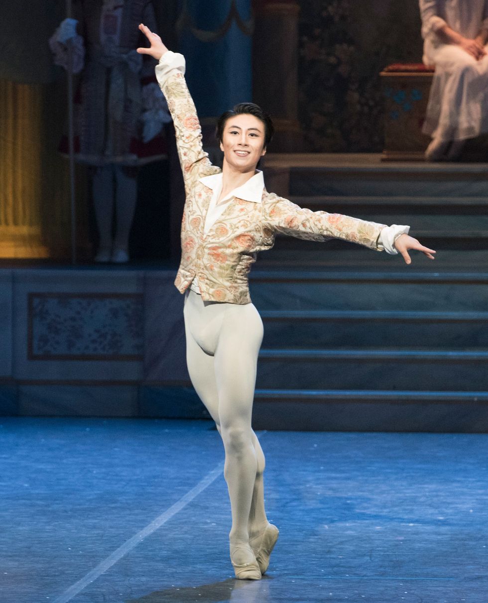 Lee stands in sous-sus onstage in white tights and a gold and white jacket, smiling at the audience.