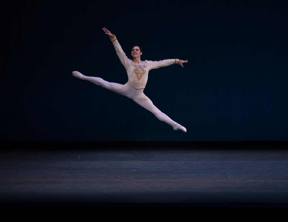 Wilkes-Davis, in white tights and a gold jacket, leaps through the air with his legs split, smiling at the audience.