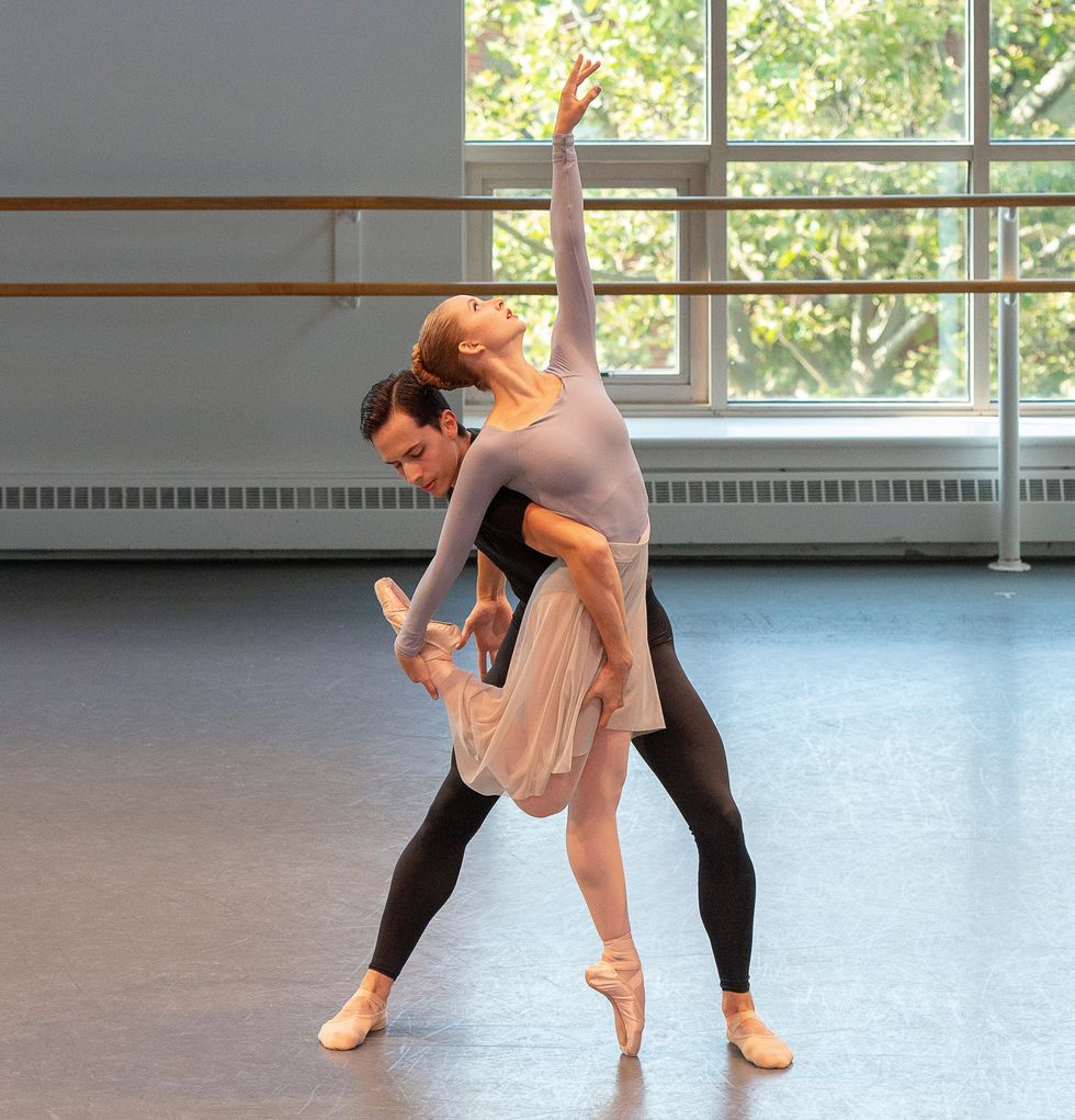 Studio shot of Novak and Wilkes-Davis in an entangled pose. Novak stands on pointe, holding one leg behind her and reaches toward the ceiling.