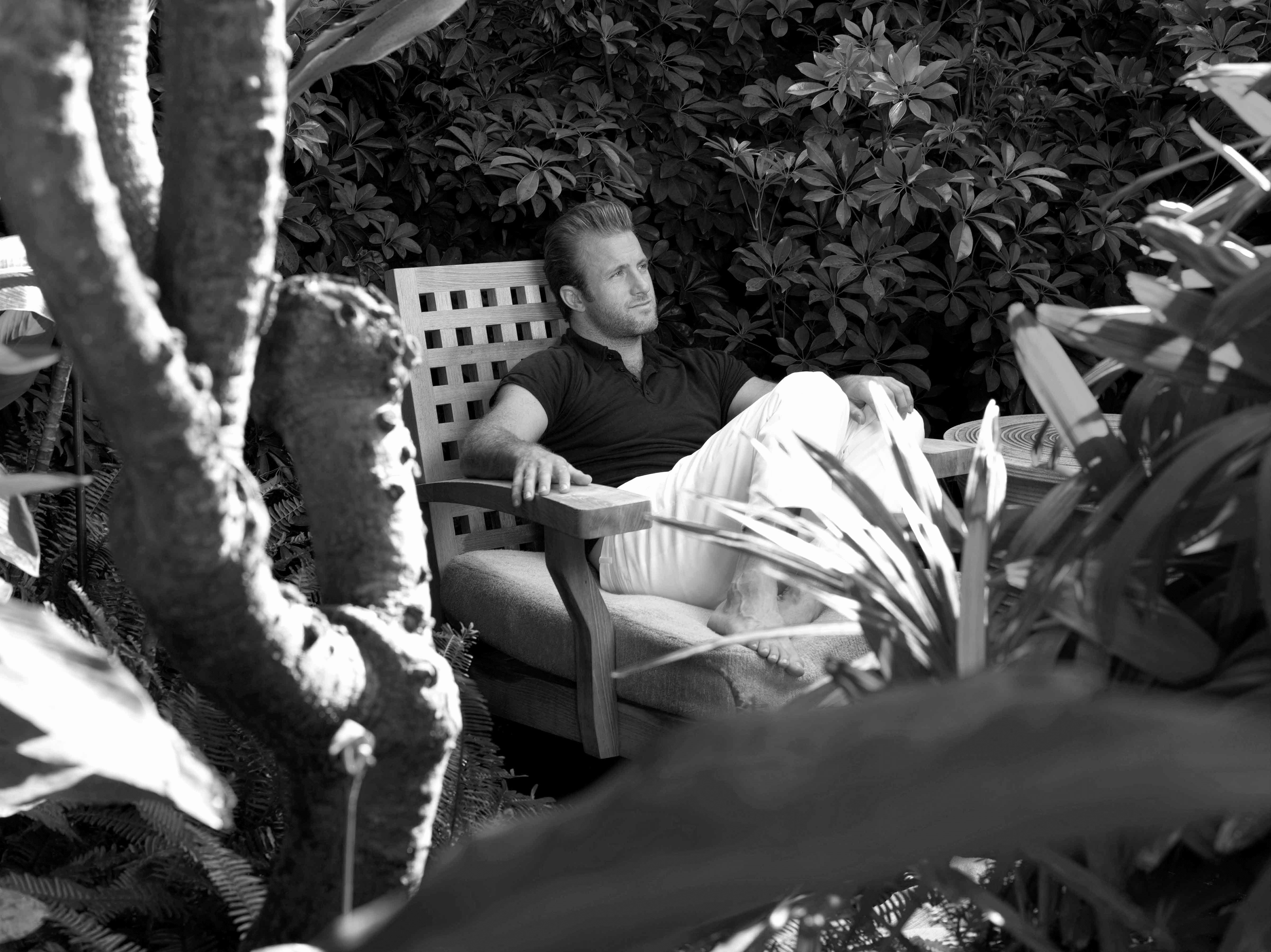 Black and white photo of Scott Caan sitting in a garden.