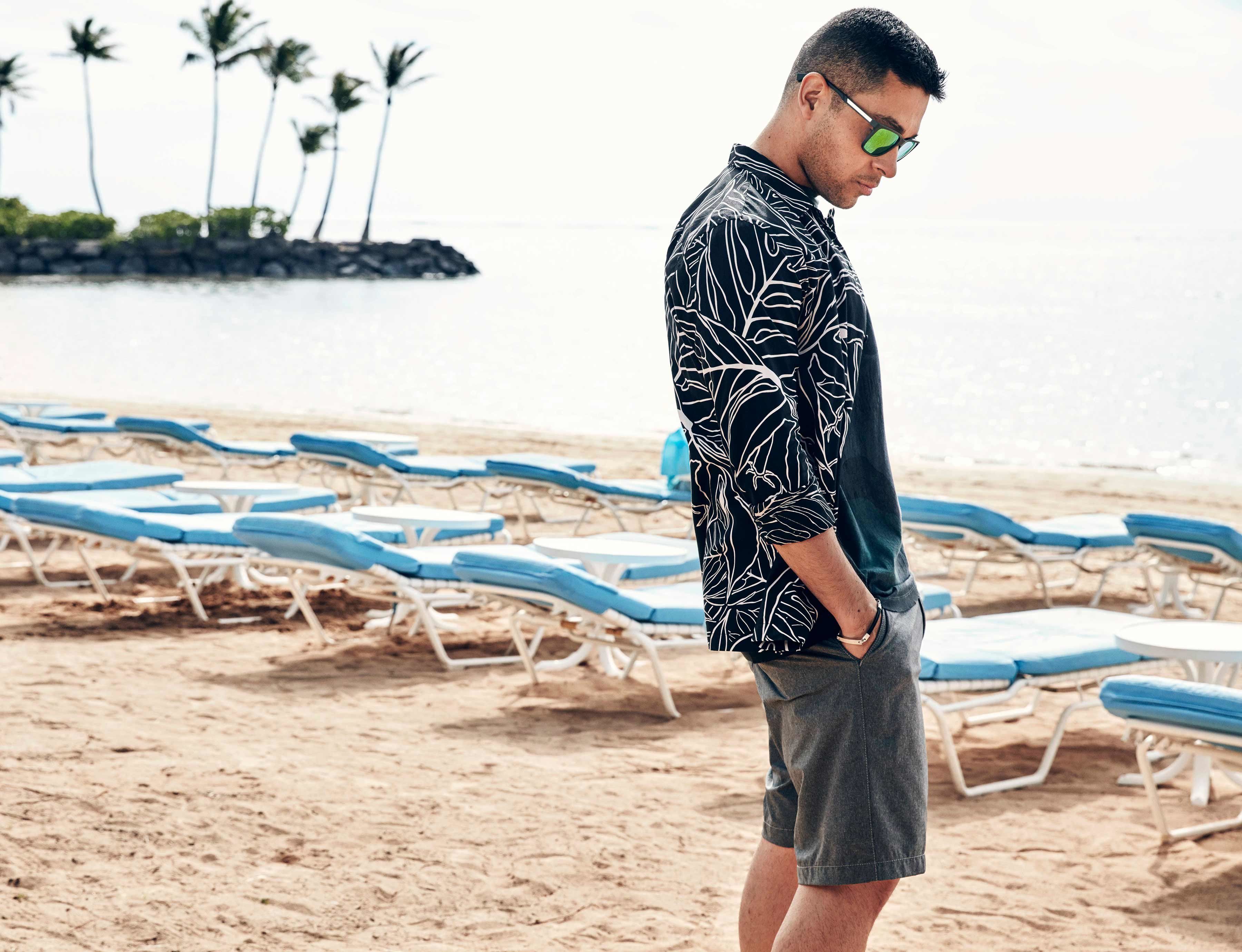 Wilmer Valderrama in a striped jacket looking down on the sandy beach in front of rows of blue lounge chairs