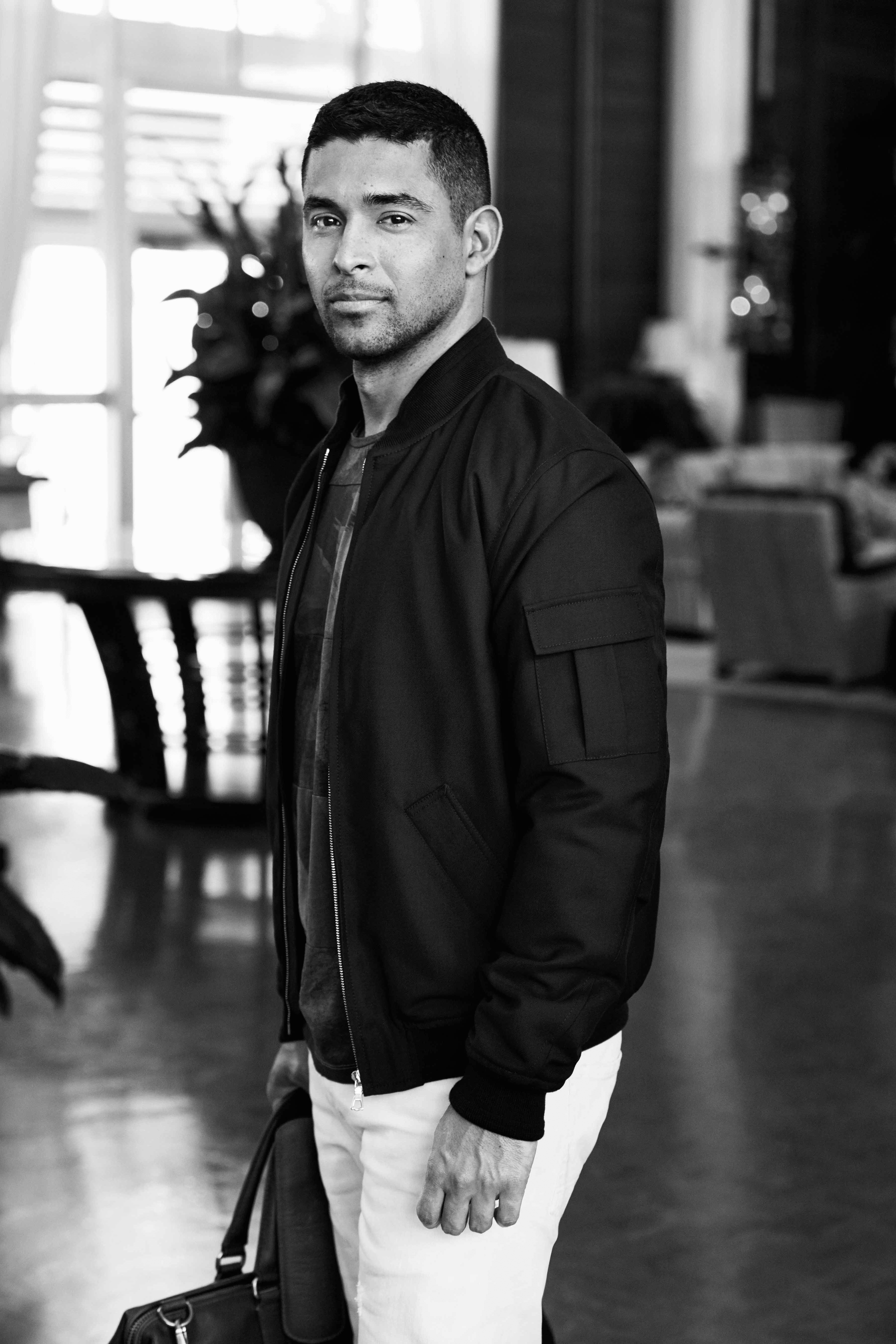 A black and white photo of Wilmer Valderrama with his body turned to the left but looking straight ahead and wearing white pants and a bomber jacket