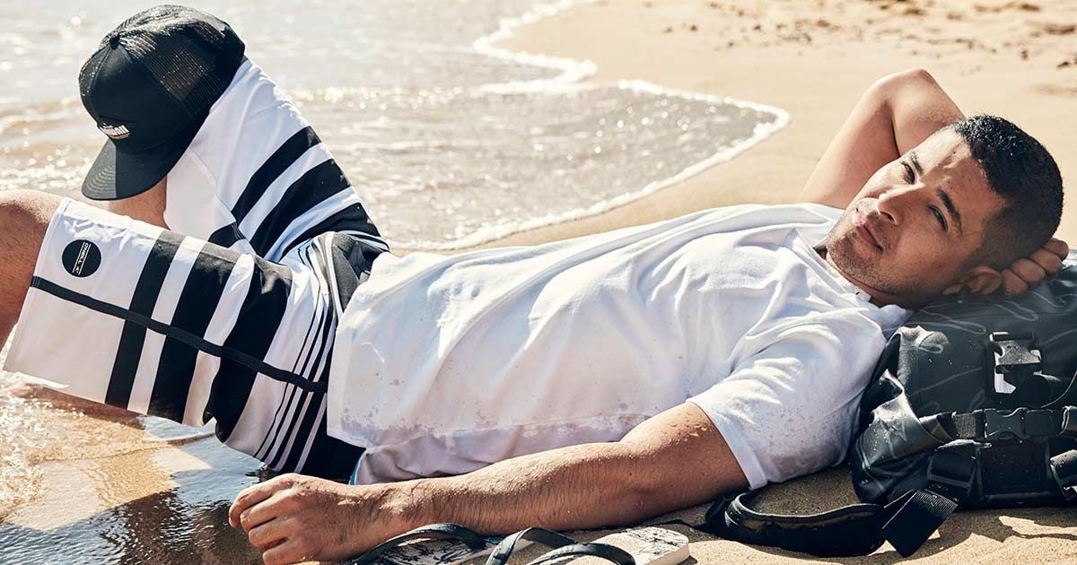 Wilmer Valderrama laying in the sand in a white shirt and white and black swim trunks using a backpack as a headrest