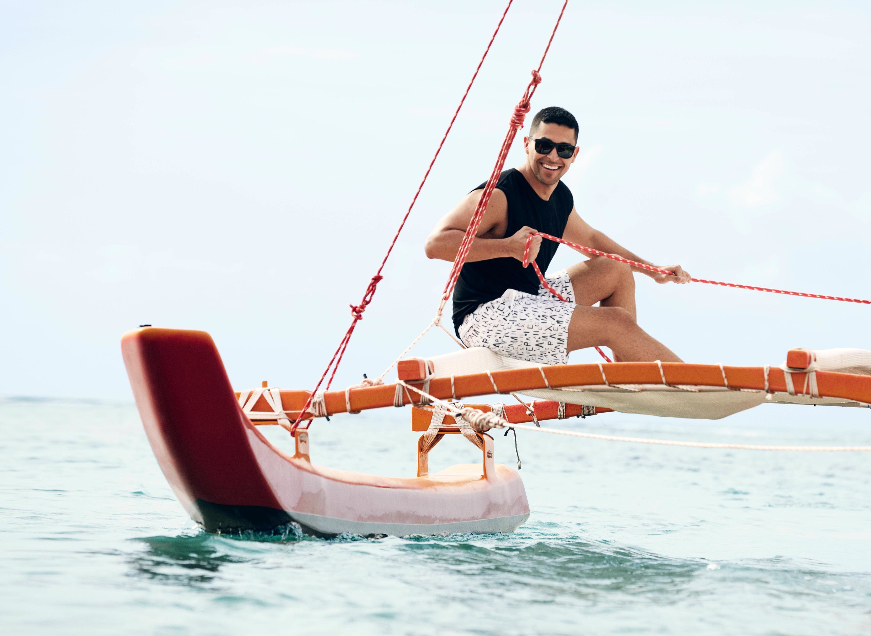 Wilmer Valderrama sailing on the ocean in a black tank top and white swim trunks