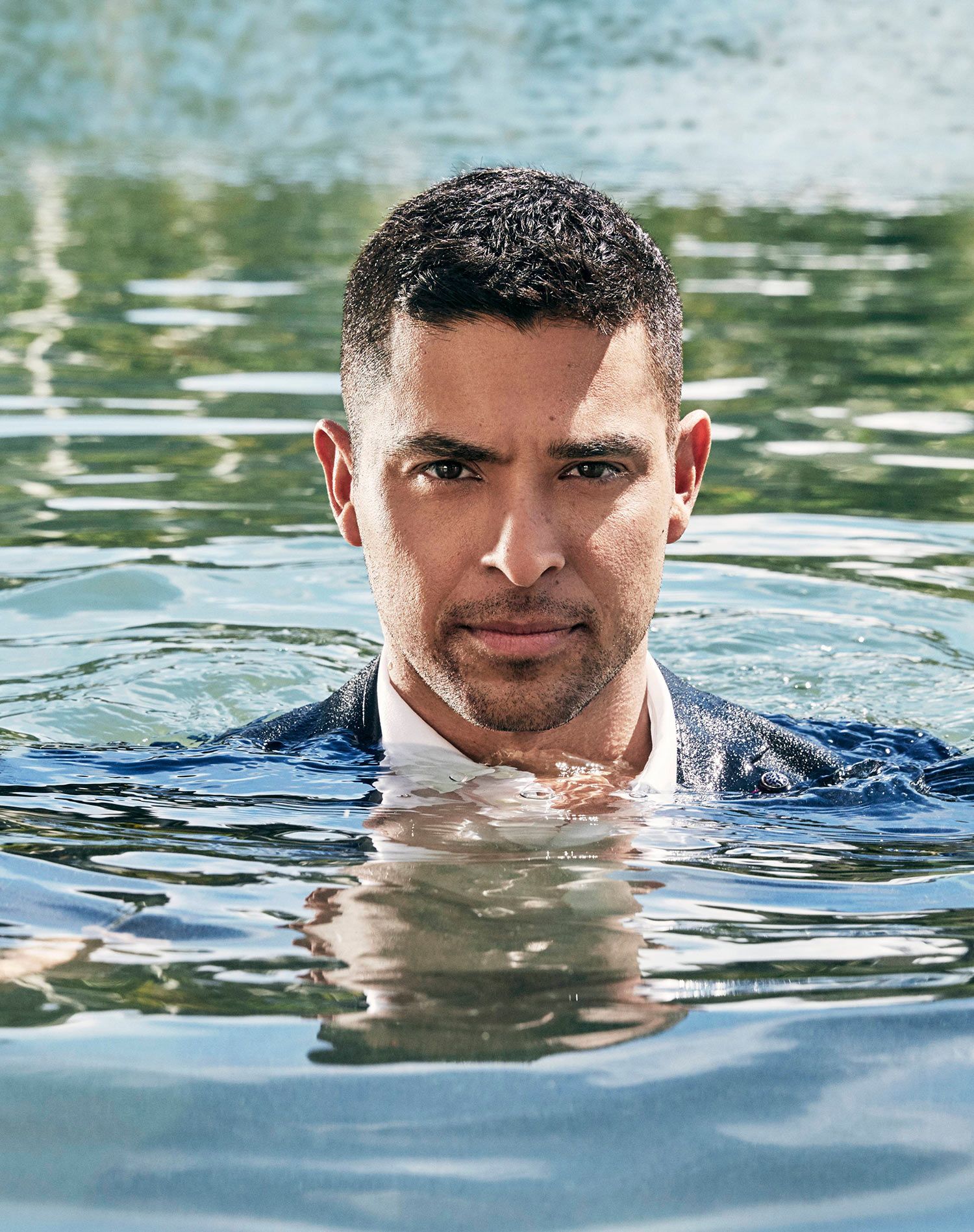 Wilmer Valderrama wearing a suit while in a body of water with just his head above the water