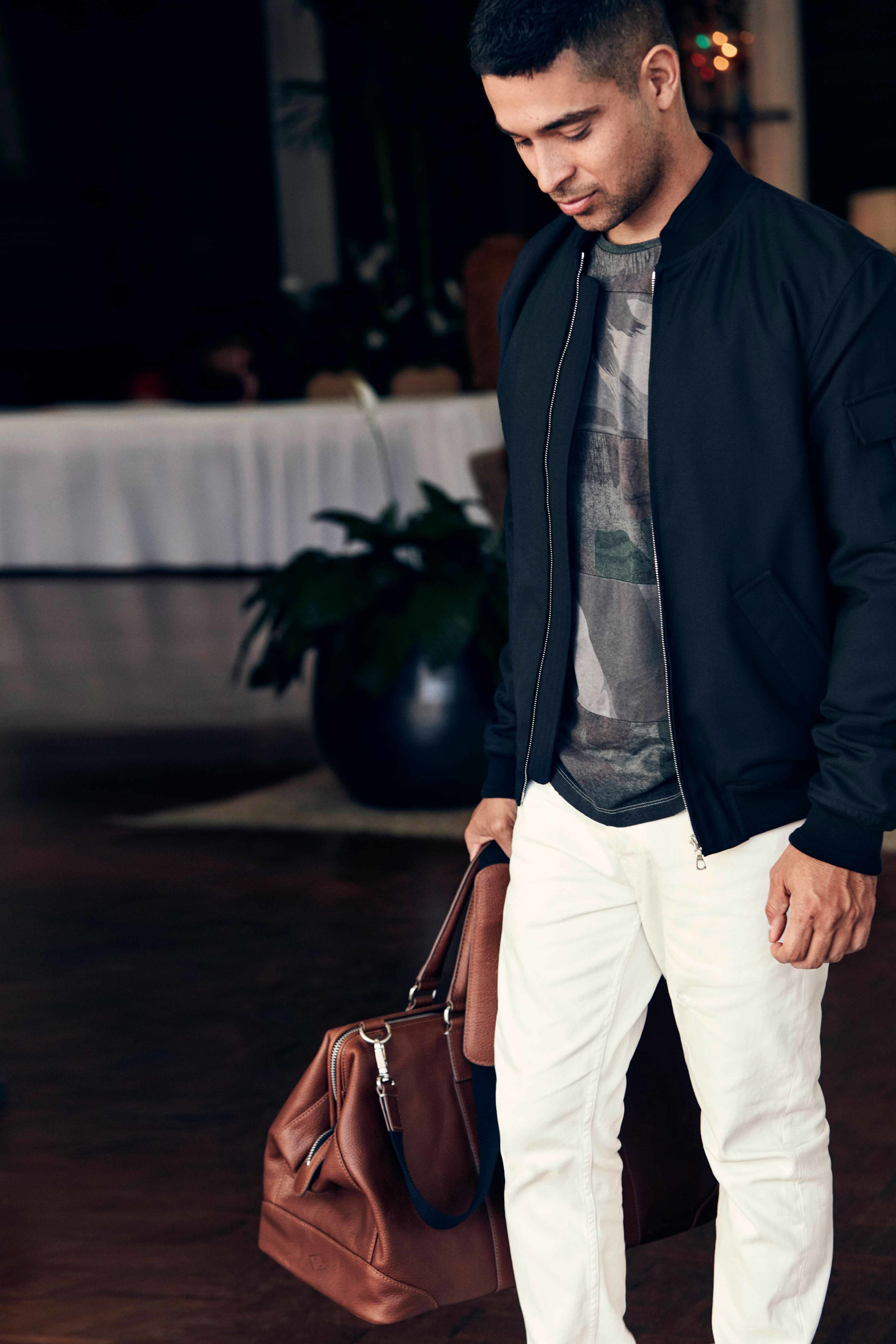 Wilmer Valderrama looking at the ground and wearing white pants and a blue bomber jacket and camouflage shirt