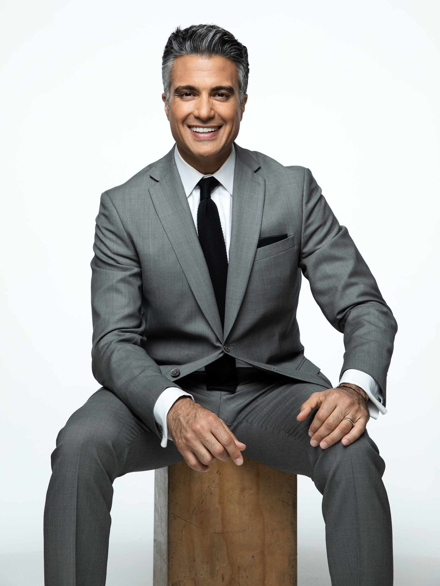 Actor Jaime Camil wearing a suit and sitting on a stool.