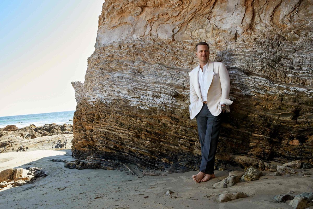 Chris O'Donnell barefoot in the sand wearing a white jacket and gray pants at the base of a cliff