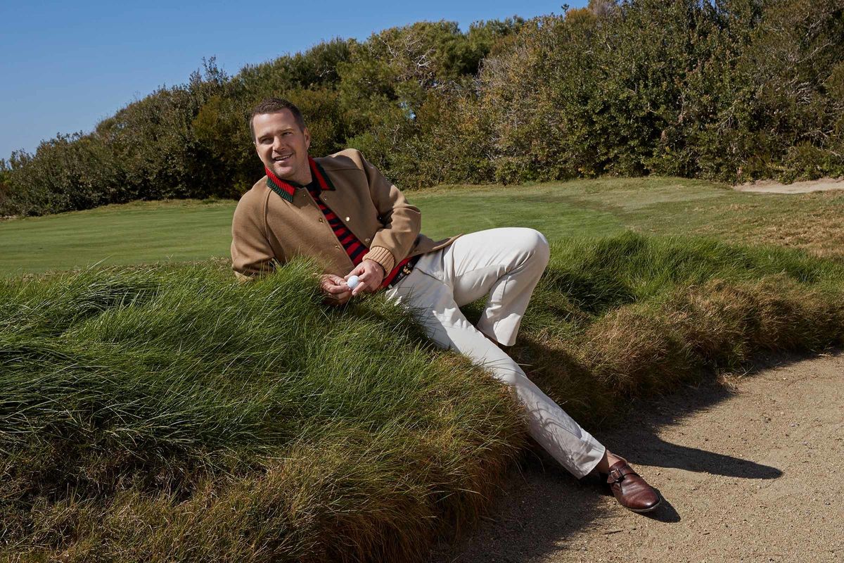 Chris O'Donnell laying in the grass while wearing a camel jacket and white pants