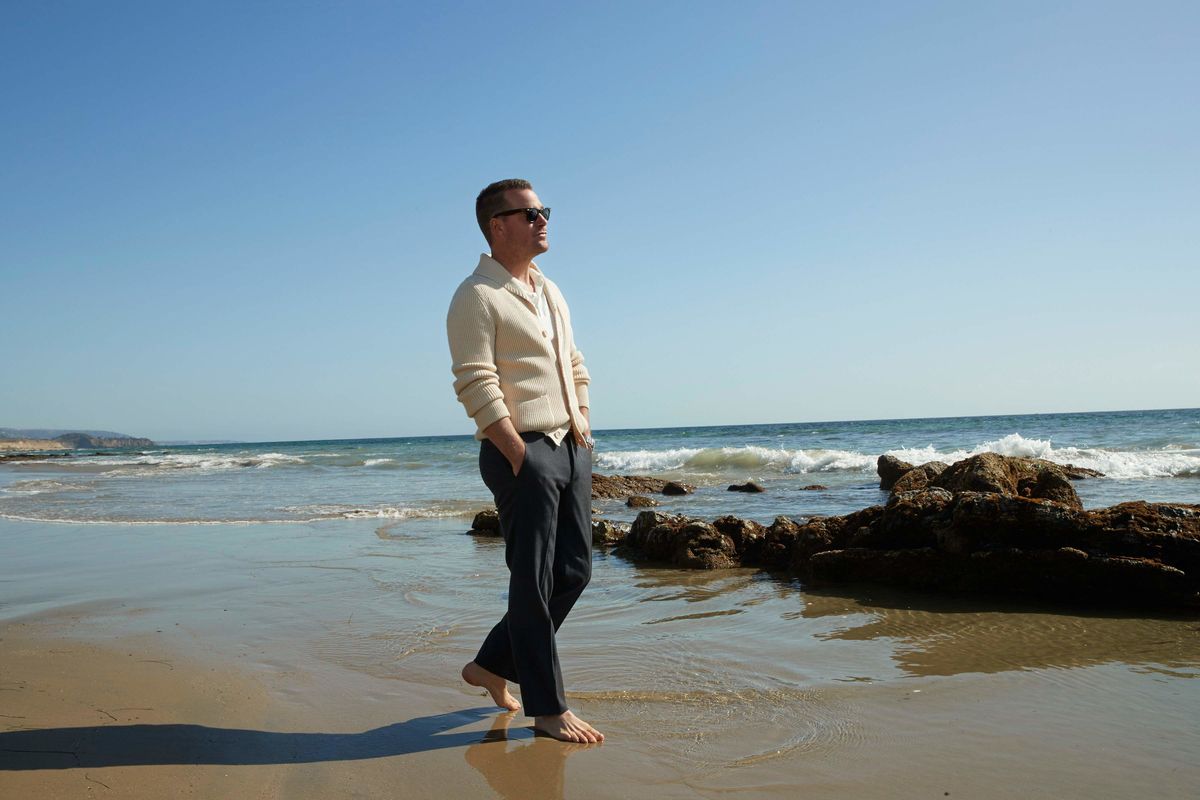 Barefoot Chris O'Donnell walking along the beach in a cream cardigan and gray pants
