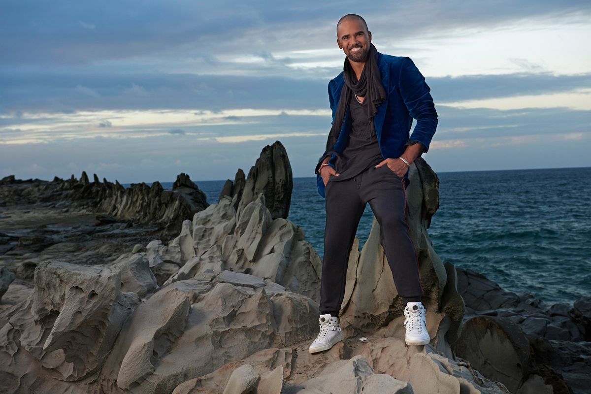 Shemar Moore standing on rocky terrain by the ocean in a velvet blue jacket