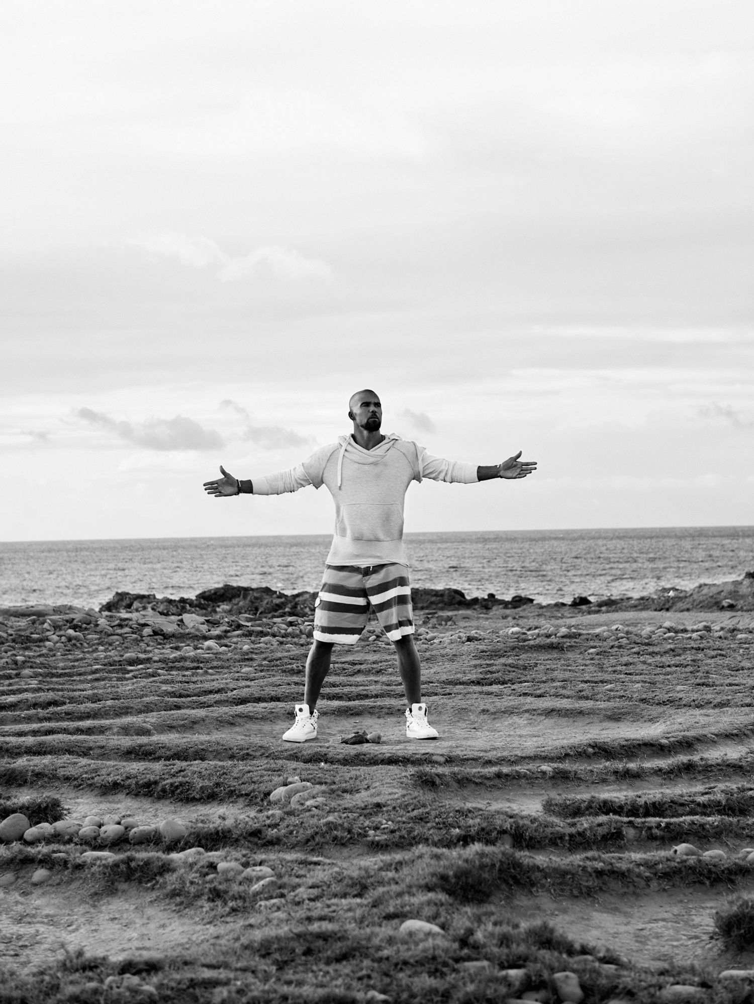 Shemar Moore in a hoodie and swim trunks standing in sand in front of the ocean