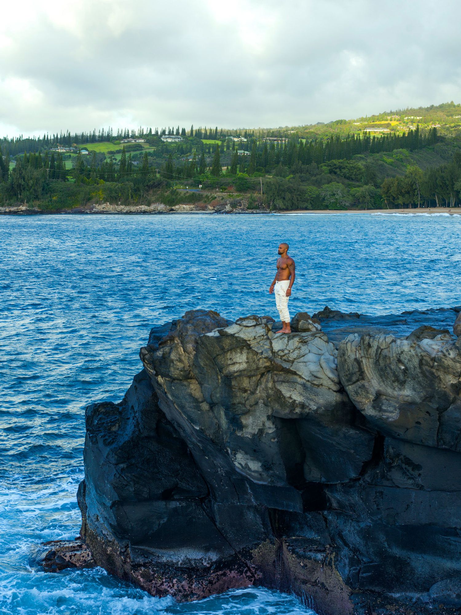 Shemar Moore standing on rocky terrain by the ocean with his shirt off and in white pants