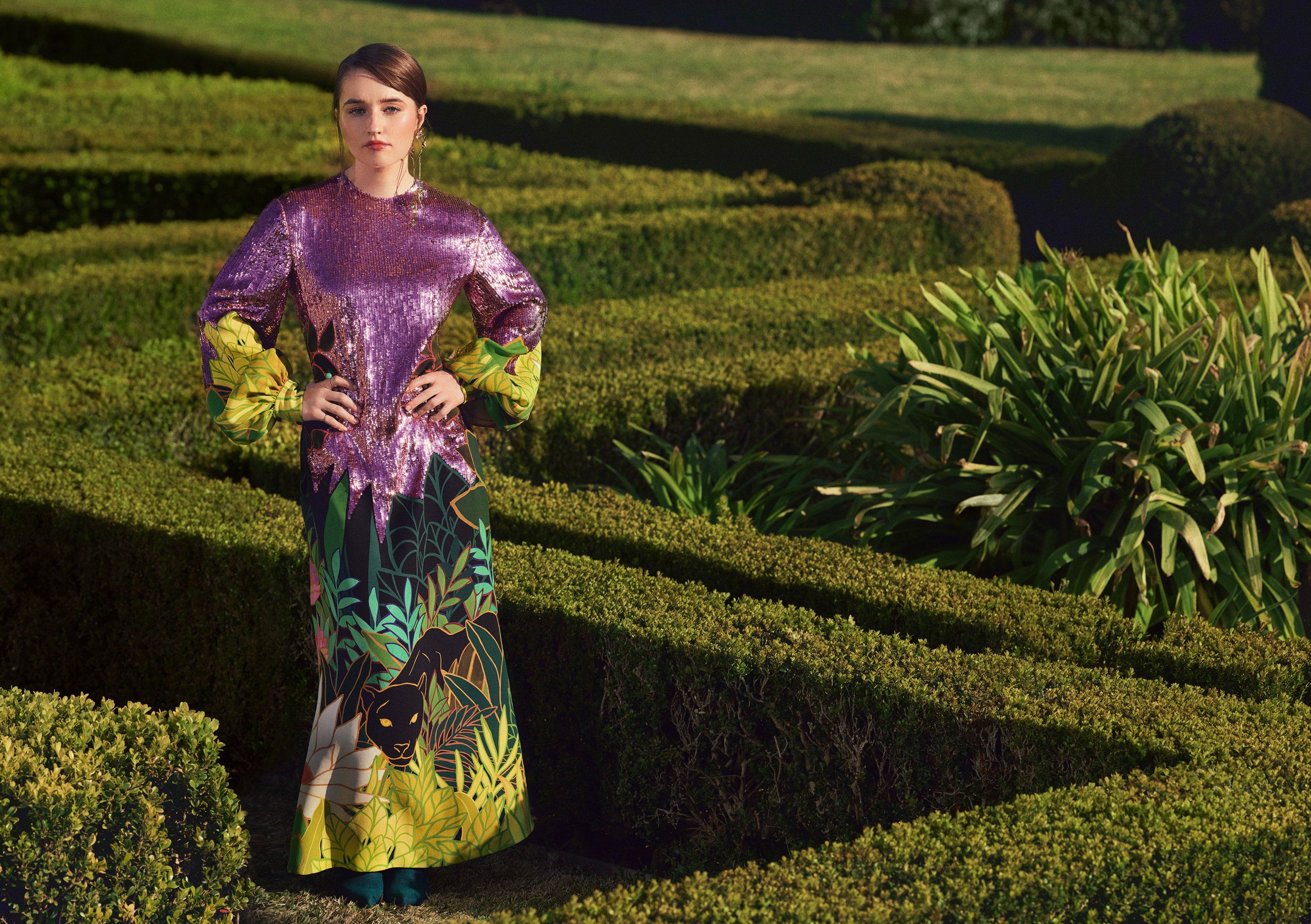 Kaitlyn Dever standing in a landscaped maze in a sequined and floral print dress