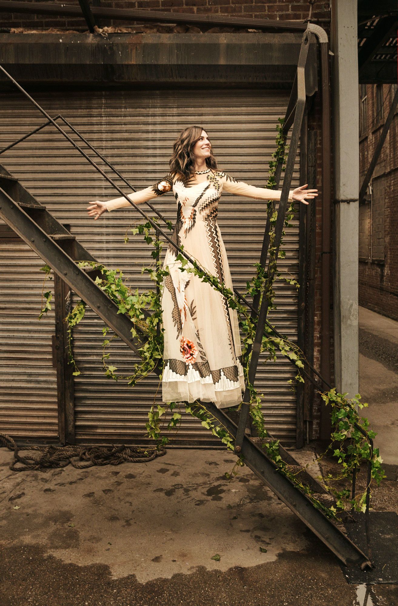 Maggie Siff standing on a fire escape in a floral dress.