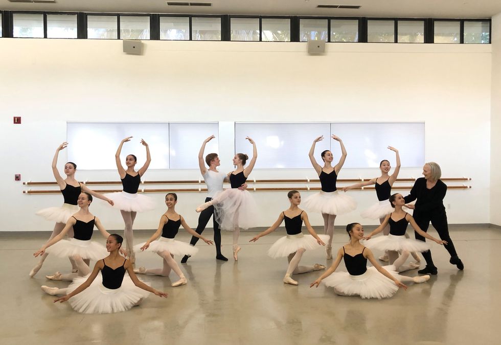 Schneider adjusts the head of young ballet student in a studio. A ballet close stands posed in rehearsal in white tutus and black leotards.